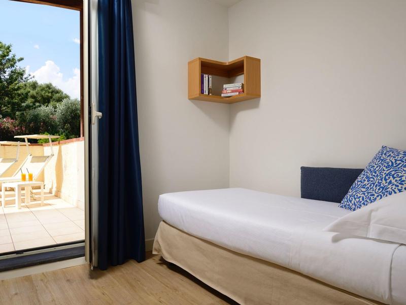 Kleine hotelkamer met eenpersoonsbed, blauw kussen, houten vloer en schuifdeur naar buitenpatio.