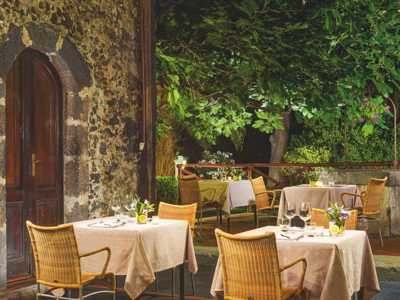 Buitenterras met tafels, stoelen en warme verlichting bij een stenen gebouw en groen.