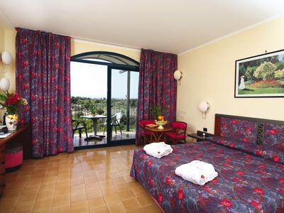 Habitación de hotel con cama doble, balcón y vista a un lago, cortinas rojas y muebles de madera.