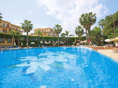 Gran piscina azul con tumbonas y palmeras frente a un hotel bajo un cielo azul.