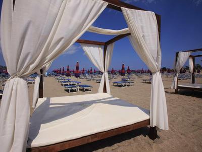 Cama con cortinas blancas en la playa con sillas y sombrillas bajo un cielo azul.