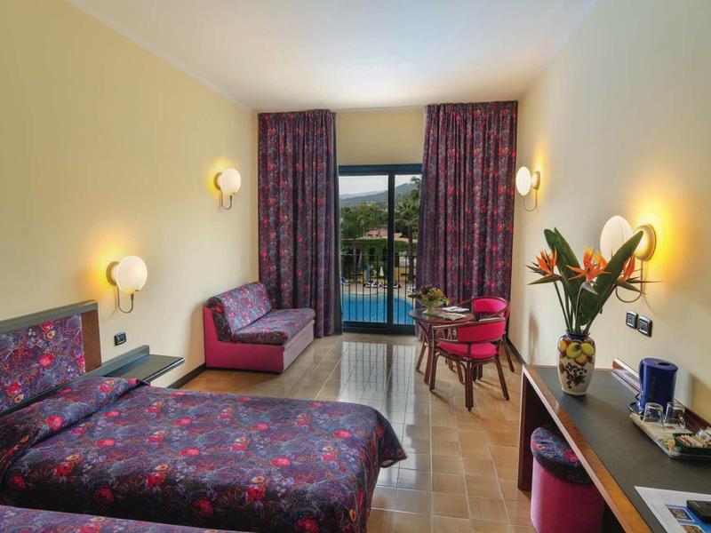 Habitación de hotel con cama doble, zona de estar, escritorio y balcón con vistas a la naturaleza.