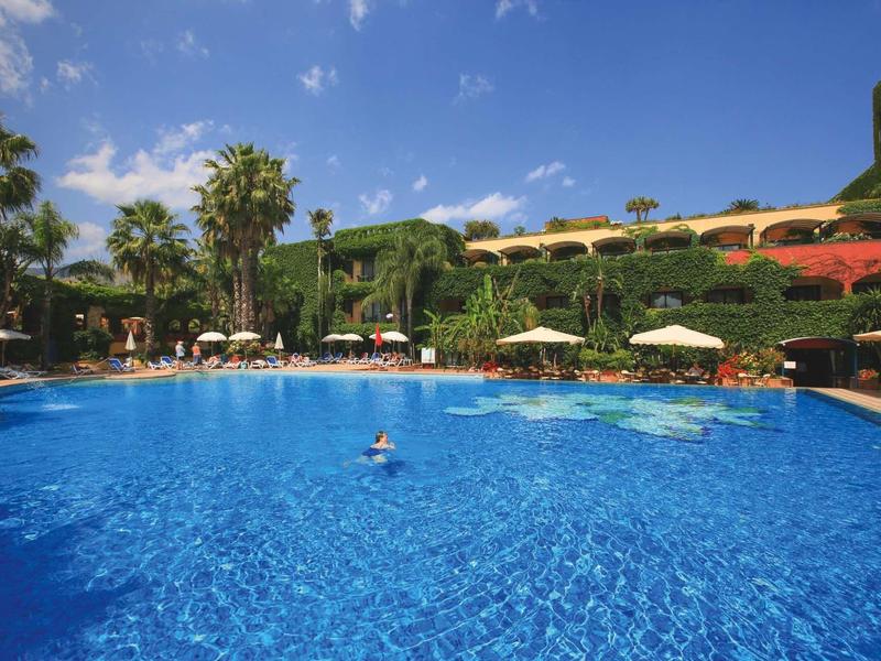Gran piscina azul de hotel con palmeras, sombrillas y huéspedes bajo cielo azul.