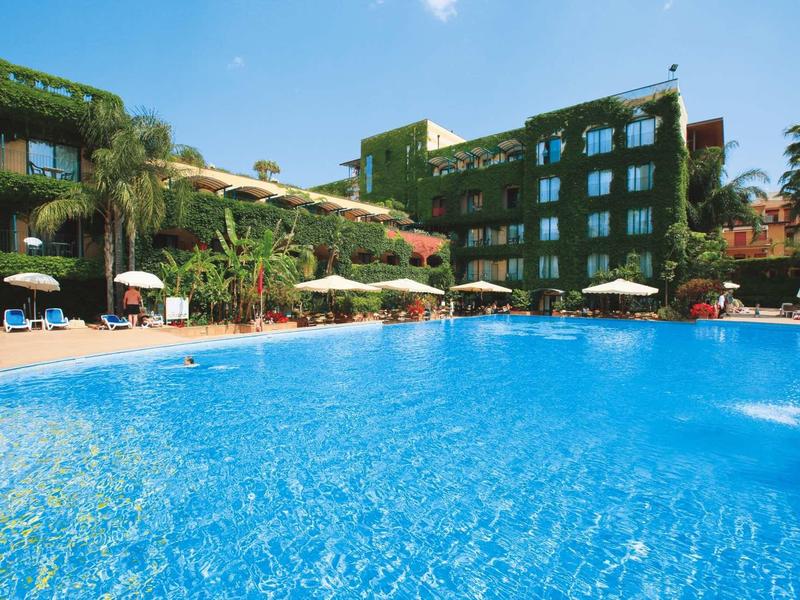 Gran piscina exterior frente a un hotel de varios pisos con plantas verdes y sombrillas.