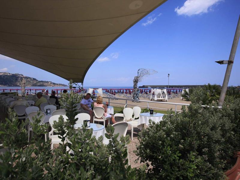 Área exterior de un café con sillas y mesas blancas bajo una vela de sombra cerca del mar.
