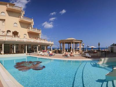 Großer Außenpool mit runder Form, umgeben von Sandsteinfarbenen Gebäuden und Sonnenterrasse.