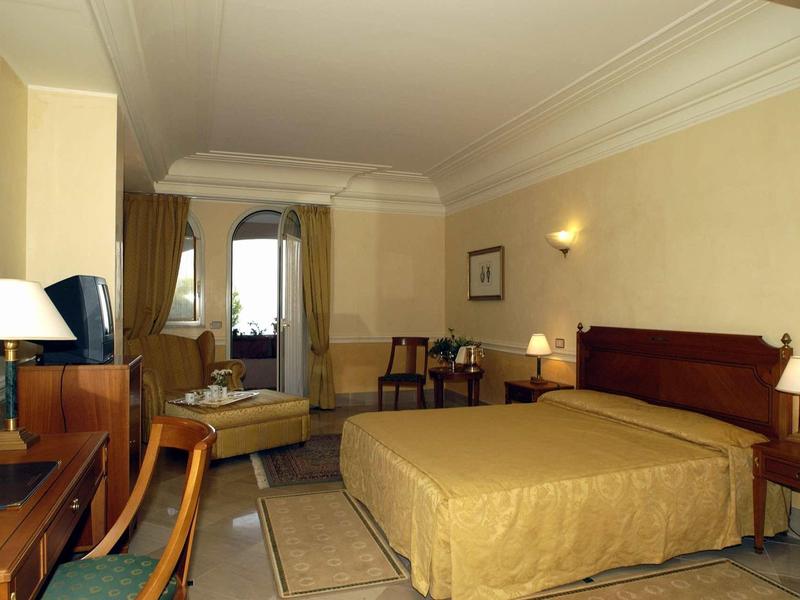 Gemütliches Hotelzimmer mit Doppelbett, Schreibtisch, Fernseher und Stuhl in warmen Beigetönen.