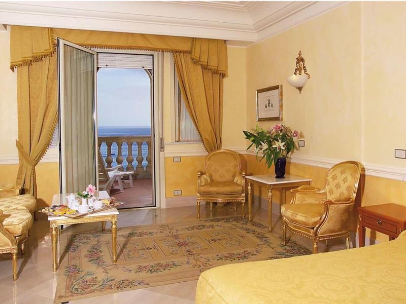 Helles Hotelzimmer mit gelbem Bett, bequemen Sesseln, Blumen und Blick auf Meer durch Balkon.