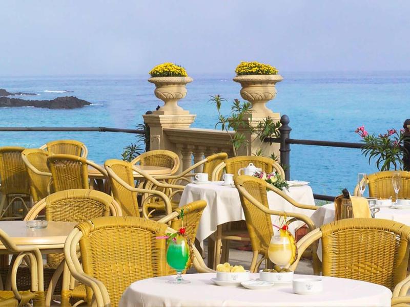 Terrassenrestaurant mit gelben Korbstühlen und weißen Tischdecken am Meer bei klarem Himmel.