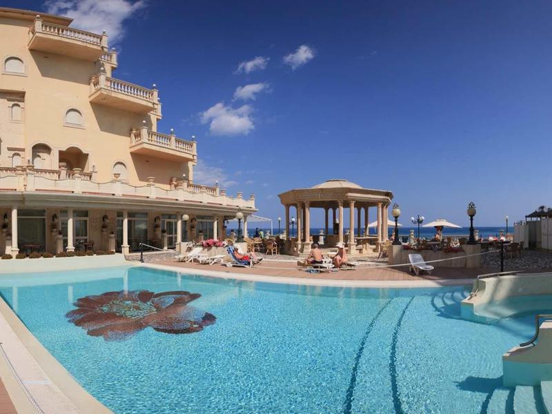 Großer Außenpool mit runder Form, umgeben von Sandsteinfarbenen Gebäuden und Sonnenterrasse.