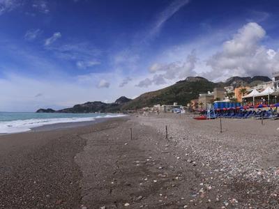 Wide beach with dark sand, umbrellas, and hillside buildings under a blue sky.