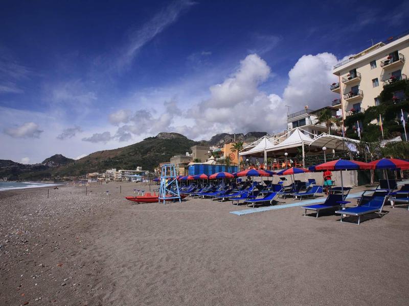 Beach with sun loungers and umbrellas in front of a hotel under blue sky.