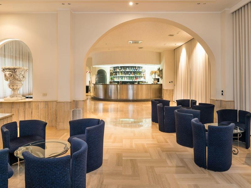 Espace lounge d'hôtel moderne avec fauteuils bleus, tables en verre et bar ouvert.