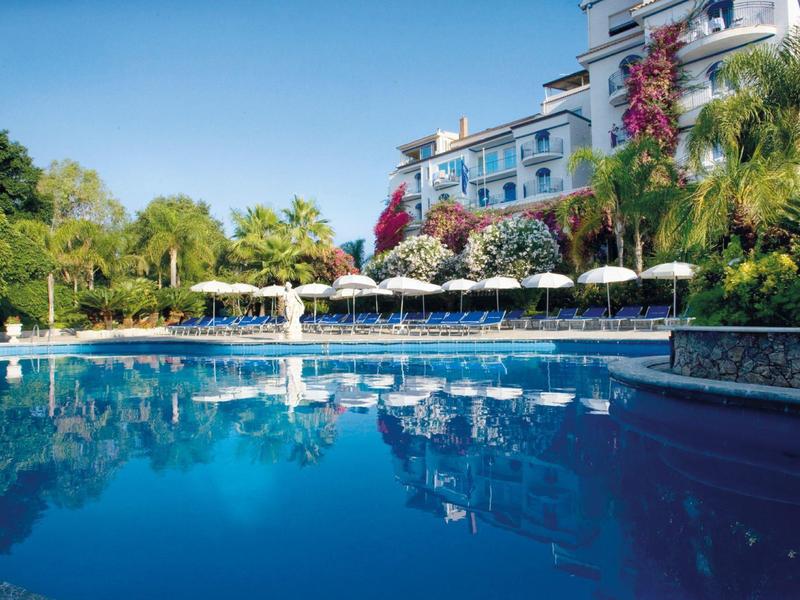 Hôtel avec grande piscine, chaises longues autour et végétation tropicale sous ciel clair.