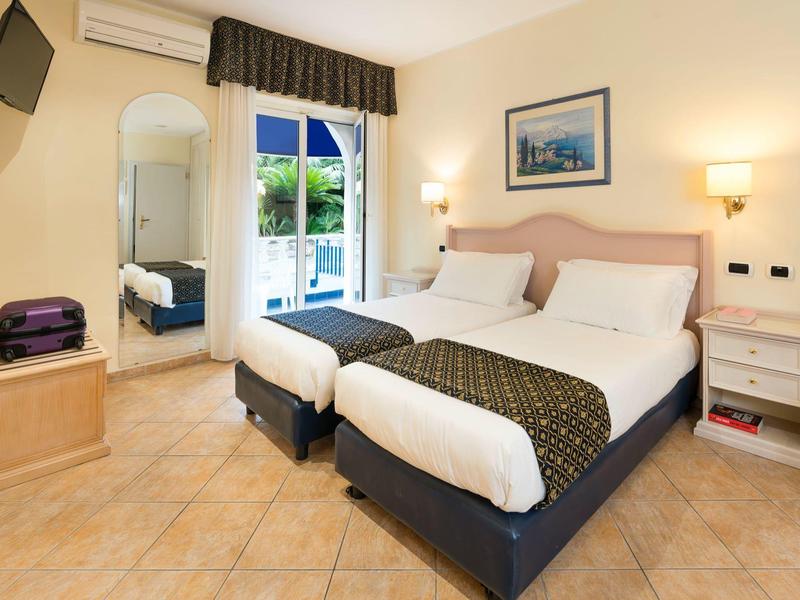 Chambre d'hôtel lumineuse avec deux lits simples, commode, miroir et porte-fenêtre.