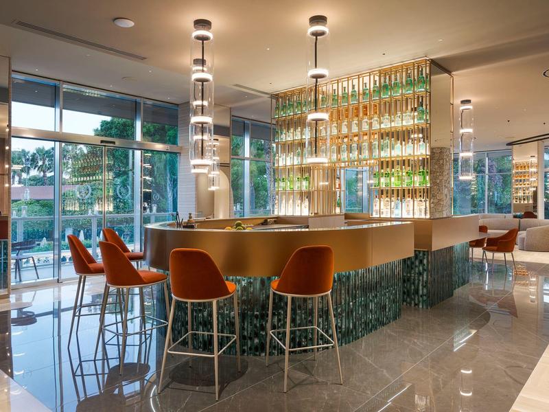 Espace bar d'hôtel moderne avec comptoir en bois, chaises orange et grandes fenêtres laissant passer la lumière du jour.