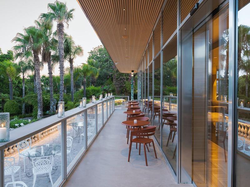 Terrasse d'hôtel moderne avec chaises et garde-corps en verre donnant sur des palmiers et un jardin.