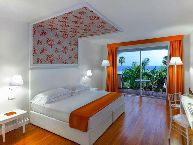 Modern hotel room with white bed, orange accents, and balcony overlooking palm trees.