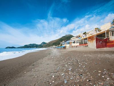Strand mit dunklem Sand und verstreuten Muscheln neben Gebäuden und Bergen unter blauem Himmel.