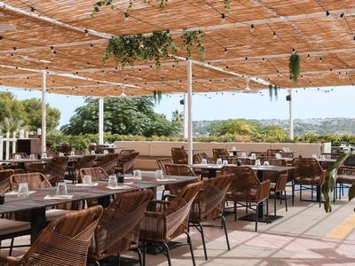 Zadaszony taras restauracyjny z stolikami, krzesłami i widokiem na zielony krajobraz.