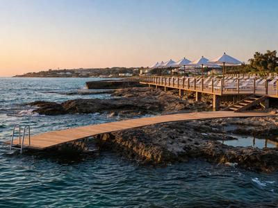 Ein Steg mit weißen Sonnenschirmen über Felsen am Meer bei Sonnenuntergang.