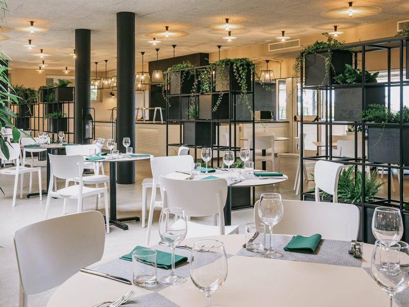 Modernes, helles Restaurant mit weißen Tischen, Stühlen und grünen Pflanzen als Dekoration.
