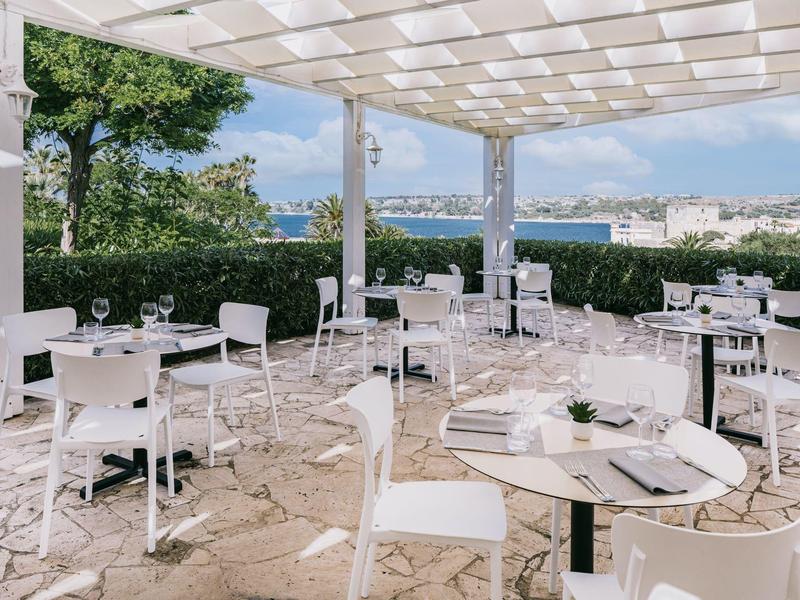 Offene Terrasse mit weißen Stühlen, Tischen, Glasgeschirr und Blick auf Stadt und Wasser.