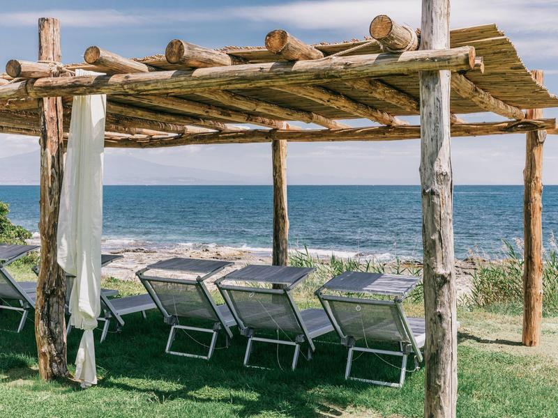 Holzpergola mit Liegestühlen auf grünem Rasen vor ruhiger, blauer Meereskulisse unter blauem Himmel.