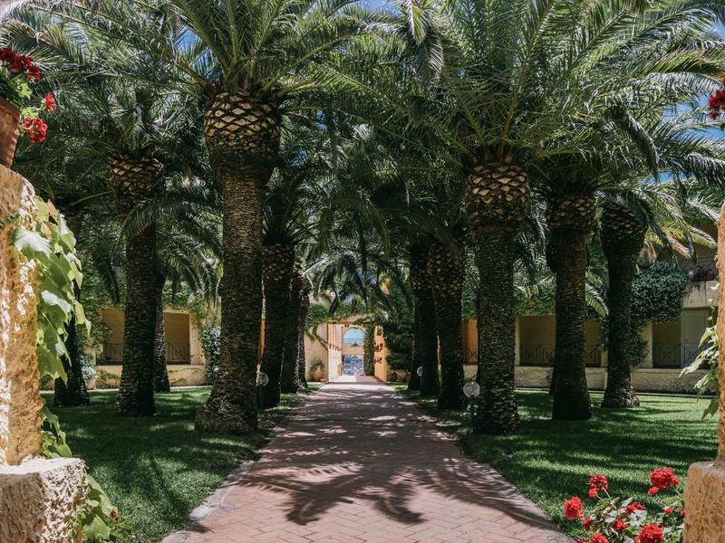 Weg gesäumt von hohen Palmen und blühenden Pflanzen führt zu einem Gebäudeeingang.