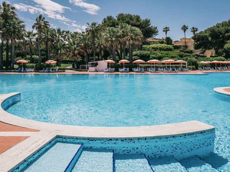 Großer blauer Pool mit mehreren Stufen, umgeben von Sonnenschirmen und Palmen unter blauem Himmel.