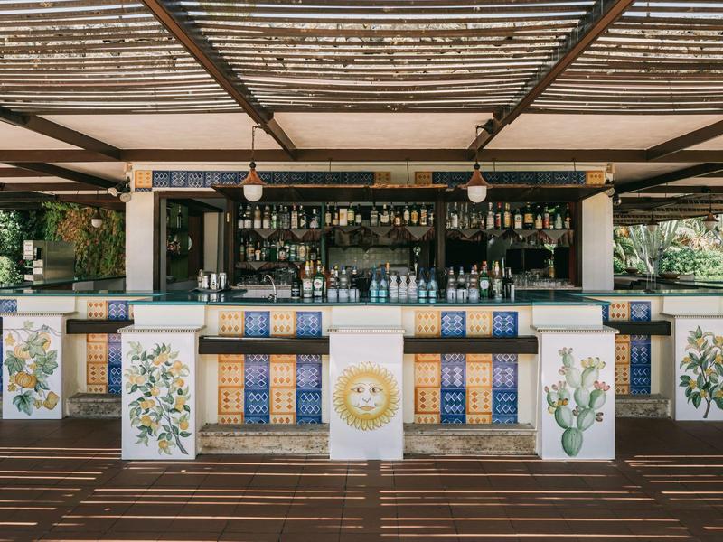 Bar mit dekorativen Kacheln, Holztheke und Regalen, überdacht mit einem Gitterdach.