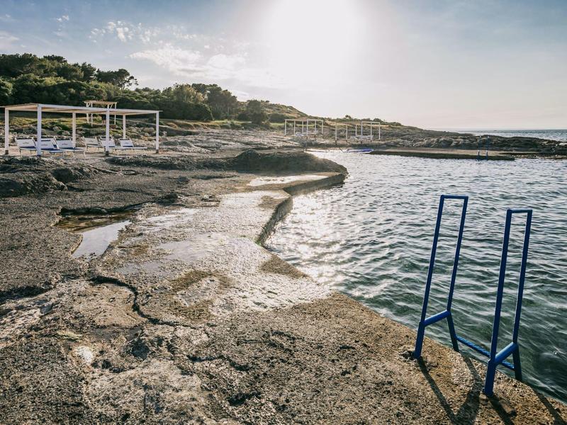 Küstenlandschaft mit steinigem Ufer, blauem Wasser und blauer Badeleiter am rechten Bildrand.