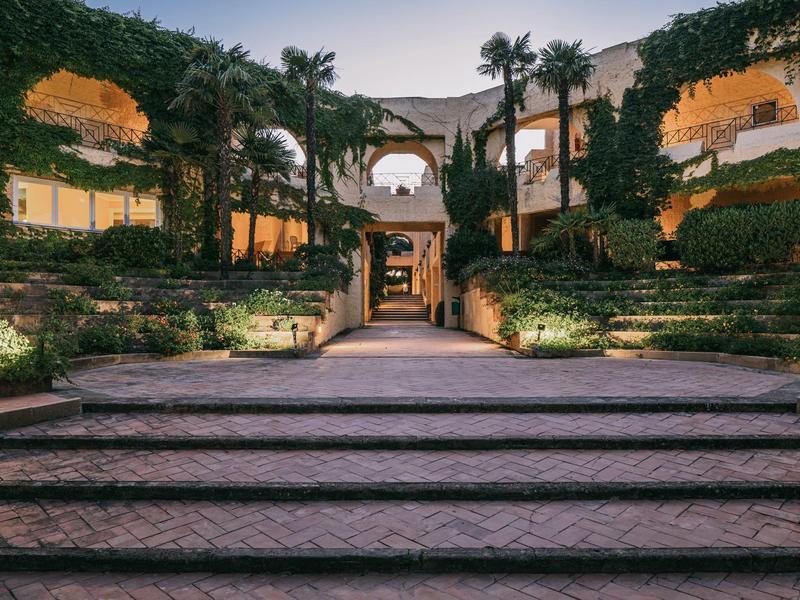 Großer Innenhof mit Treppe, begrünten Mauern, Palmen und warmen Lichtern bei Abenddämmerung.