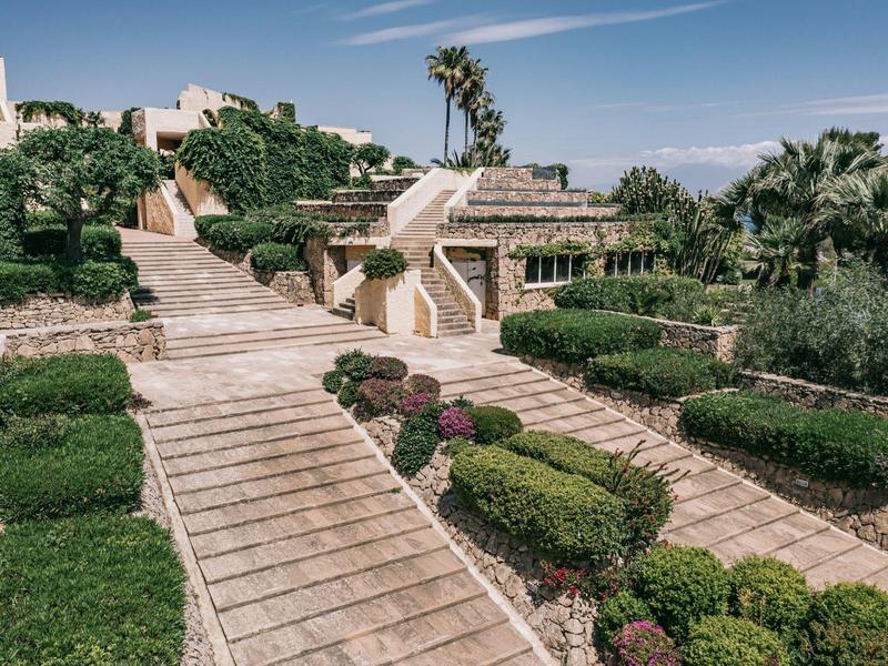 Großer Außenbereich mit breiten Treppen, grünen Hecken und Palmen unter blauem Himmel.