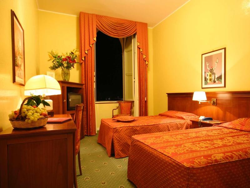 Pokój hotelowy z dwoma pojedynczymi łóżkami, ciepłym oświetleniem i czerwonymi zasłonami przy oknie.