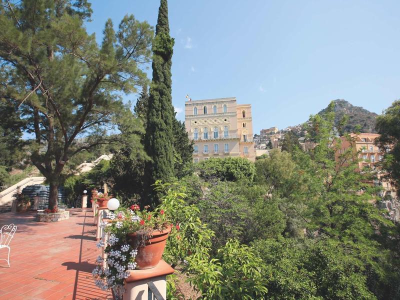 Taras z czerwonymi płytkami, dekoracjami kwiatowymi i widokiem na duży budynek i zielone drzewa.