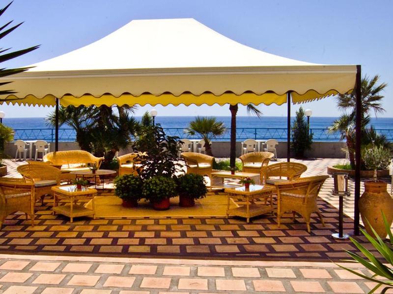 Zadaszony teren na zewnątrz z stołami i krzesłami, widok na morze i palmy w tle.