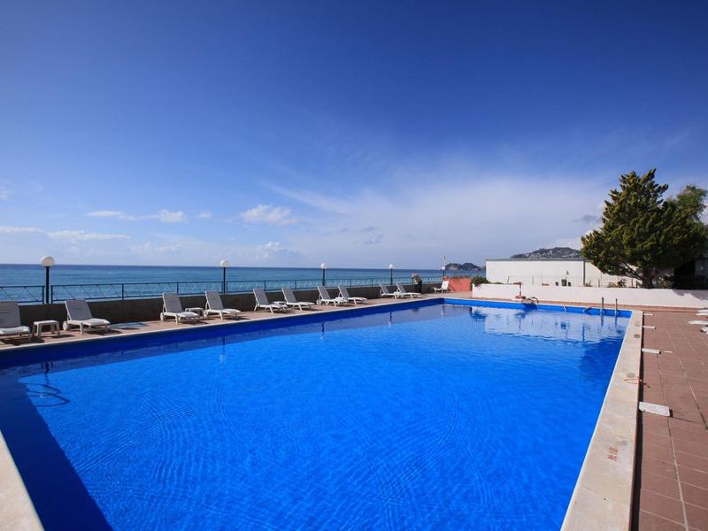 Duży basen zewnętrzny z leżakami i widokiem na morze w pogodny dzień.