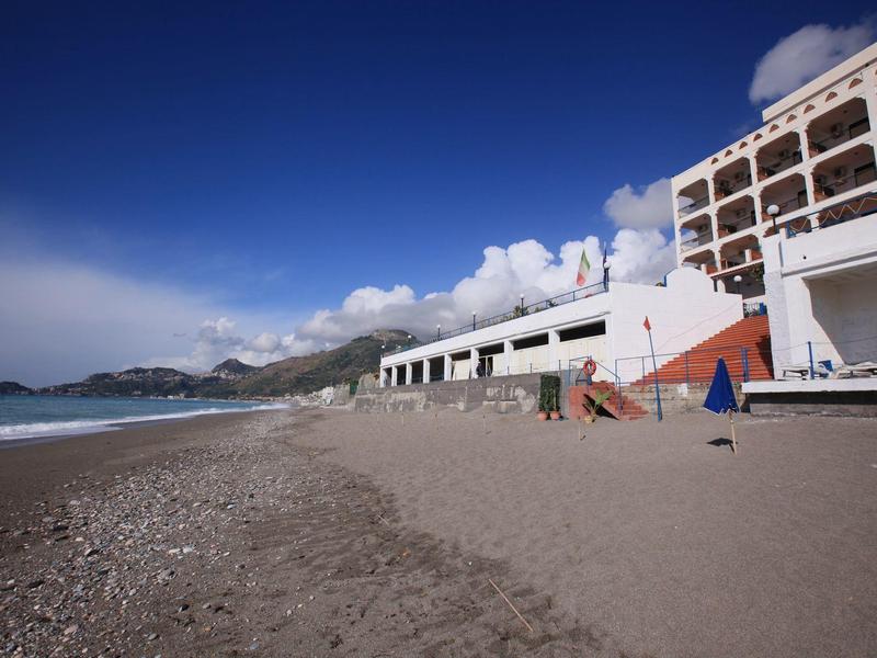 Szeroka piaszczysta plaża z białym budynkiem hotelu pod jasnym niebieskim niebem.