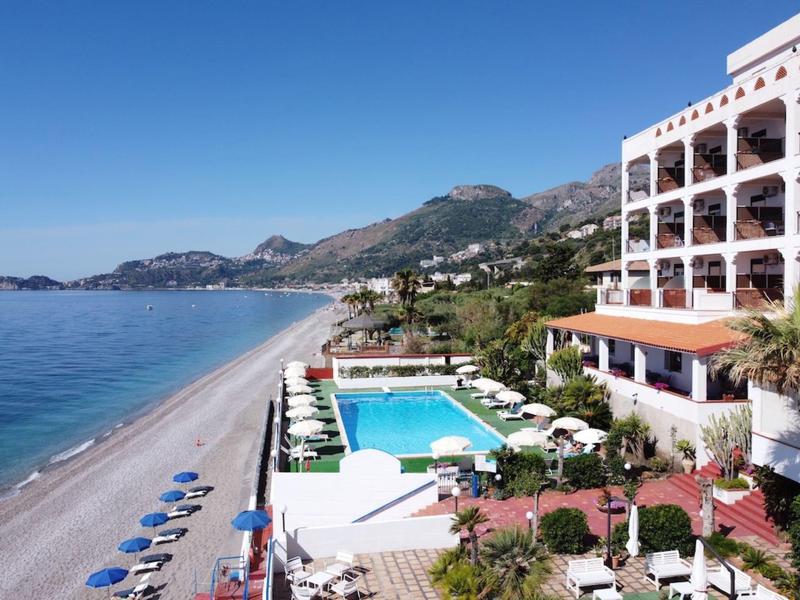 Hotel nadmorski z basenem, leżakami i widokiem na długą piaszczystą plażę i góry.