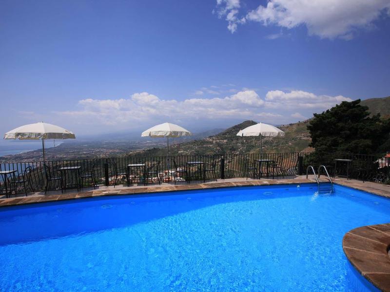 Blauw zwembad met witte parasols en bergzicht onder een blauwe lucht.