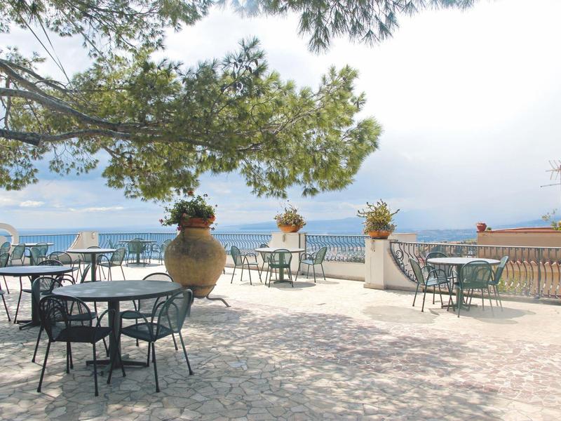 Terrace with metal chairs and tables, decorated with large pots, overlooking the sea under trees.