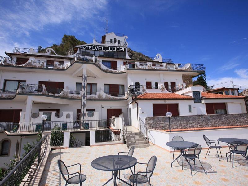 Multi-story hotel with balconies and outdoor terrace with tables and chairs under blue sky.