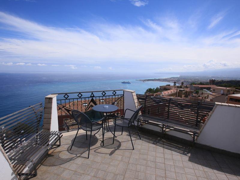 Balcony with chairs and table overlooking sea and city under blue sky.
