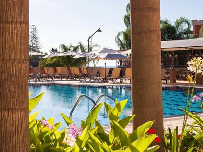 Zwembadgebied met ligstoelen en parasols tussen palmbomen en tropische planten.