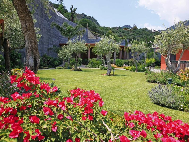 Grüner Garten mit roten Blumen, Bäumen, Rasenfläche und Bergen im Hintergrund unter blauem Himmel.