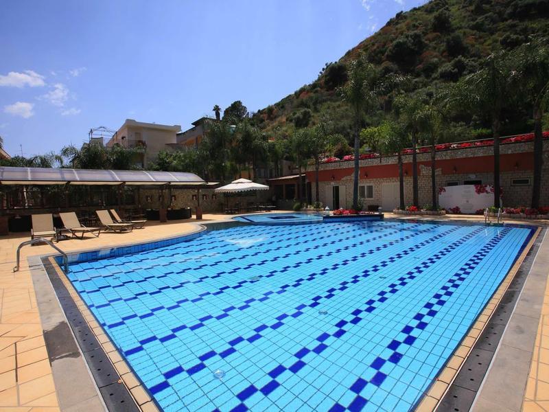 Rechteckiges Schwimmbecken mit blauen Fliesen und Liegestühlen neben einem bewaldeten Hügel.