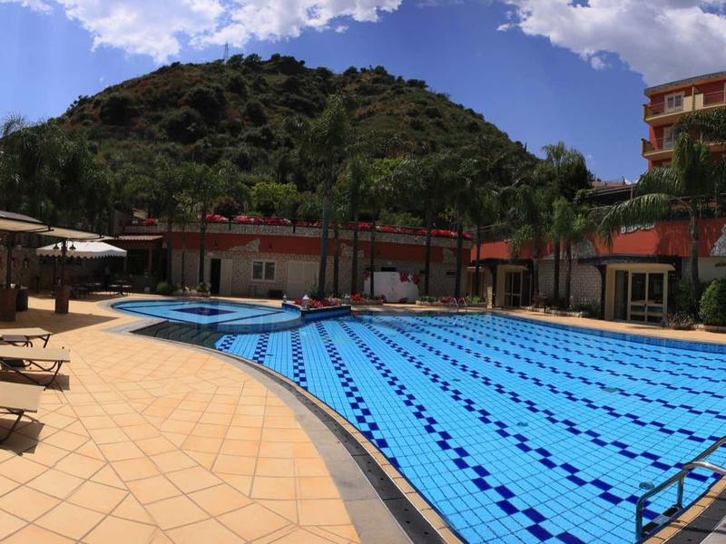 Großes Außen-Schwimmbecken mit Bahnen, Liegestühlen und Hügeln im Hintergrund bei Sonnenschein.