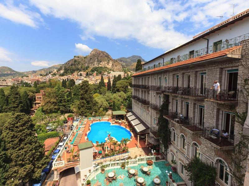 View of a hotel with a swimming pool, terrace, and mountains in the background.