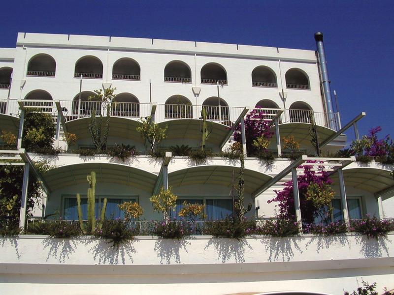 Meerverdiepingen wit hotelgebouw met balkons en bloeiende planten onder een heldere blauwe lucht.